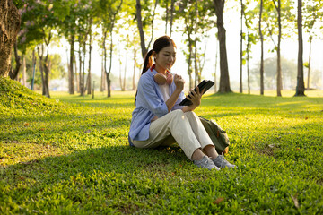 A woman is sitting on the grass in a park, listening to music. She is wearing headphones and has a book in her lap. Concept of relaxation and leisure
