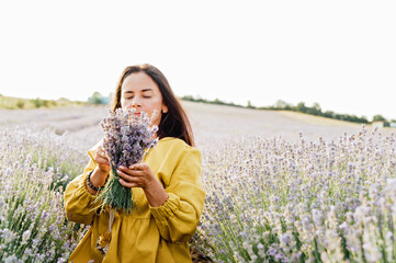 woman in yellow dress stands in lavender field, gently holding bouquet of lavender flowers. She appears serene and contemplative, enjoying beauty and fragrance of blossoms around her.