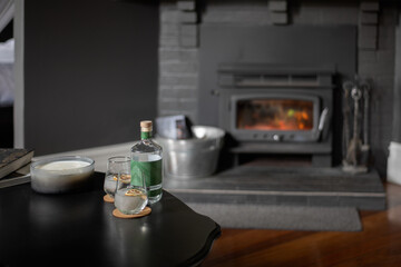 a bottle of gin with two glasses, and 2 gin and tonic cocktails in front of a fire