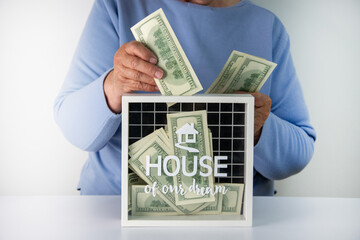 A person is putting dollar bills into a money box that says House of our dream on it.