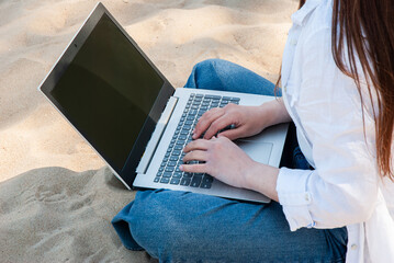 Young adult female using a laptop on a sandy beach wearing a white shirt and jeans, illustrating...