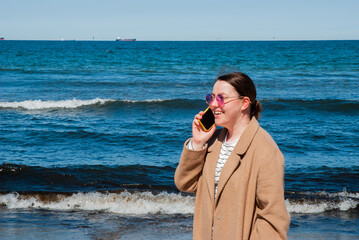 Young caucasian woman in a brown coat talking on a smartphone at the beach with blue ocean waves in the background on a sunny day. Digital nomad lifestyle. Remote work