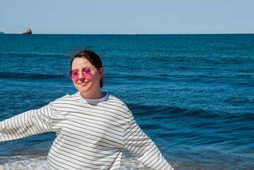 Fototapeta premium Young caucasian female with striped sweater and sunglasses enjoying a sunny day at the beach with vibrant blue ocean waves creating a serene backdrop. Digital nomad lifestyle. Remote work