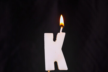 close up on a white letter K birthday candle with fire on a black background.
