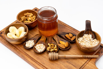 Almonds,walnut,oats and sliced banana kept in bowls and spoons with a jar of honey on a wooden board