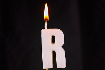 close up on a white letter R birthday candle with fire on a black background.
