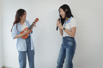 Two happy young Asian girls holding microphone and ukulele while they standing in living room. Cheerful teenage women enjoy singing and playing music together. People with music and instrument.