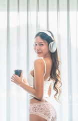 Naklejka premium Beautiful Asian Gen Z woman in loungewear enjoying coffee and music at home. Relaxed morning moment with headphones and warm drink in bright bedroom.
