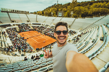 Handsome young tennis spectator taking selfie picture watching grand slam tournament match at the stadium - Sport and technology life style concept