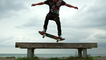 Young skater making professional trick at cloudy nature. Sporty hipster training extreme skating jumping from bench at sea. Active guy performing skate kickflip at embankment in slow motion