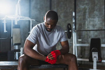 Fitness workout in gym. African American man fighter preparing for fight wrapping hands with red...