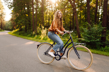 Obraz premium A joyful woman with curly hair rides a bicycle along a path in a sunny park. Young woman having fun and enjoying a day off outdoors. Active lifestyle.