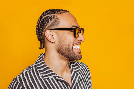 Side view portrait of a cheerful young multiracial man with braids wearing glasses, smiling and showing teeth against a vibrant yellow background.
