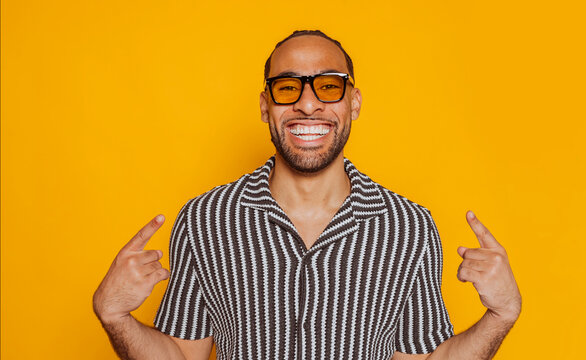 Happy young mixed race man pointing at himself wearing yellow sunglasses and smiling on a yellow background.