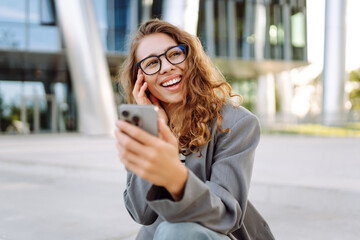Beautiful woman in stylish clothes and with phone sitting on office building background. Young...