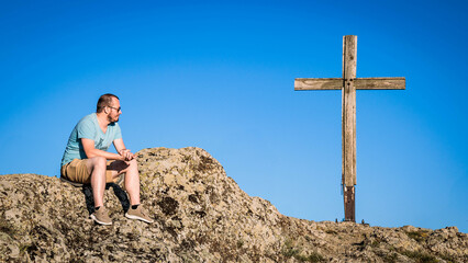 Man sitting on a rocky mountain peak near a wooden cross under clear blue sky, reflecting and...