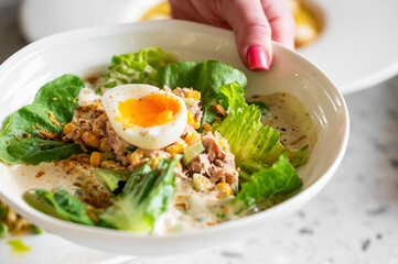 A close-up of a fresh salad with lettuce, tuna, corn, and a soft-boiled egg, garnished with seasoning and dressing. A hand with red nail polish holds the bowl. The image is vibrant and appetizing