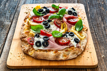 Pinsa Romana with mozzarella and burrata cheese, boiled ham, artichoke, cherry tomatoes, black olives and basil leaves on wooden table
