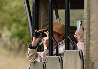 People sitting in specialized open safari vehicle, looks through binoculars and filmed on mobile phone. Travel in savannah, observe wild animals in natural habitat. Kruger national park, South Africa © Sea_Inside_Soul
