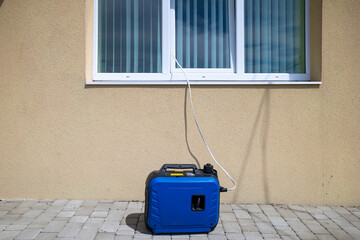 Portable blue generator on a paved surface, connected via a power cable entering through a window with vertical blinds.Temporary solution to the power supply problem.