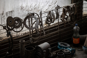 Various greasy metal parts hang from a wire mesh against a dirty window, with more tools and components scattered on a workbench below. A cardboard box sits to the left.
