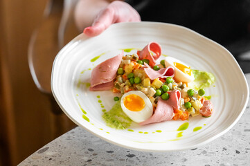 Gourmet dish with sliced ham, soft-boiled eggs, green peas, and a fresh vegetable salad, garnished with a vibrant green sauce, beautifully presented on a white plate.