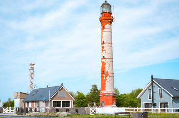 Rustic red-and-white Pakri lighthouse in Estonia stands tall against a bright blue sky, surrounded by small cozy buildings and lush greenery