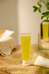 Vertical photo of a tube containing essence extracted from fresh lemon. Another white tube of cream is placed on a rattan carpet. Behind it is a table mirror reflecting the images of objects.