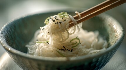 Fototapeta premium Delicate rice noodles with sesame seeds and herbs in a textured bowl, lifted by chopsticks, showcase an artful presentation of a simple yet flavorful dish.