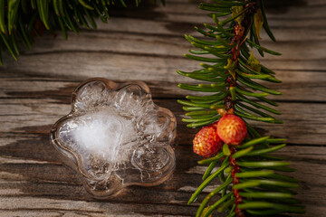 A close-up of a fir branch with new red cones paired with a clear ice paw print shape resting on rustic wooden boards.