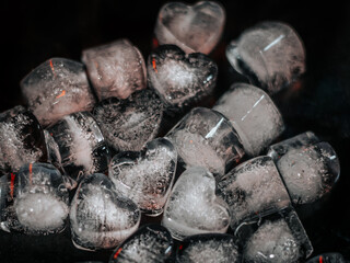 Close-up of heart-shaped and square ice cubes on a dark surface with intricate frozen textures and faint red highlights.