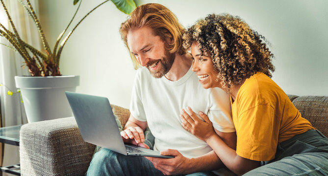 Multiracial young couple watching computer laptop sitting on the sofa at home - Happy diverse husband and wife using pc online services - Technology life style concept