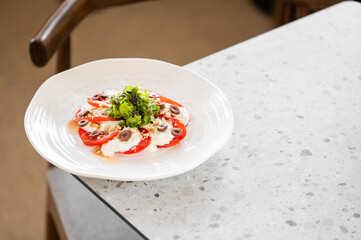 Fresh salad with sliced tomatoes, creamy cheese, olives, and greens on a white plate placed on a speckled countertop. Colorful and appetizing dish, ideal for healthy eating, culinary presentations