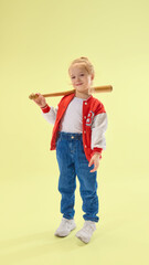 Cheerful girl holds bat on shoulder, smiling confidently, in red jacket and denim pants over yellow background. Concept of childhood, sport, activity, game, fun, leisure