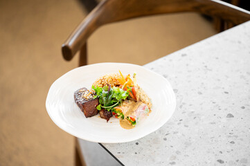 A gourmet dish featuring seared meat garnished with sesame seeds, accompanied by rice, fresh greens, and vegetables wrapped in rice paper with a creamy sauce, elegantly served on a white plate.