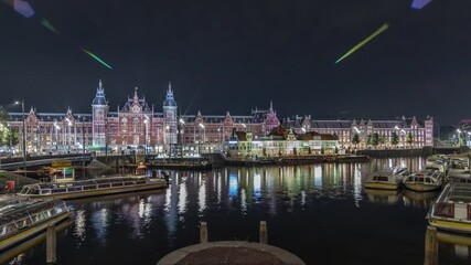 Panorama showing illuminated Central Railway station night timelapse. Tour boats moored to a jetty, trams and canal in front of the railway station in Amsterdam, The Netherlands