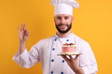 Happy confectioner in uniform holding cake with berries and showing ok gesture on orange background