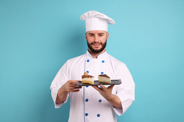 Happy confectioner in uniform holding pieces of delicious cheesecake on light blue background