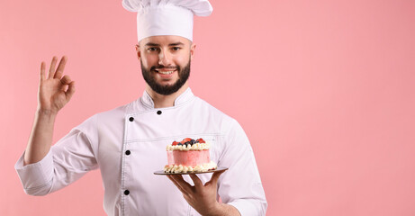 Happy confectioner in uniform holding cake with berries and showing ok gesture on pink background