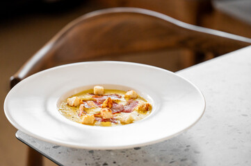 A creamy gourmet soup garnished with crispy croutons and delicate slices of prosciutto, served in a white bowl on a marble table. Elegant presentation, perfect for fine dining and culinary photography