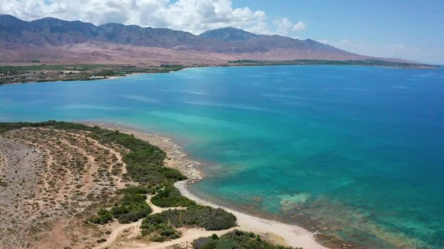 A stunning aerial shot of Assyk Kul Lake, featuring crystal-clear blueish-green waters with high mountain peaks in the background and lush greenery surrounding the lake.