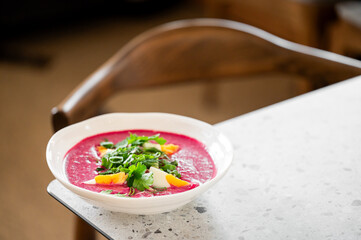 A bowl of vibrant beetroot soup garnished with fresh herbs and slices of boiled egg, placed on a speckled countertop with a wooden chair in the background