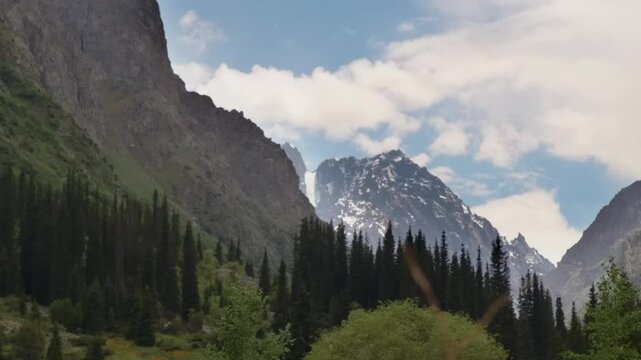 This footage captures a beautiful green valley nestled between green mountains with snowy peaks, within Ala Archa National Park, Kyrgyzstan.