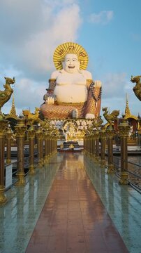 Giant laughing Buddha statue at Wat Plai Laem in Koh Samui, Thailand, surrounded by ornate golden pillars and reflecting in the temple tiled walkway. Vertical orientation