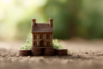 Miniature house sits atop stacked coins, nestled in greenery against a blurred natural background, symbolizing homeownership, savings, and financial stability