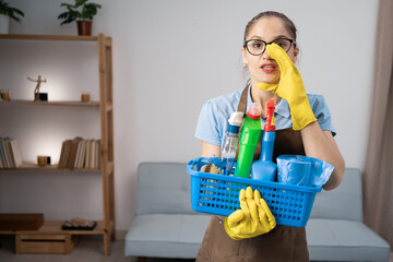 Female janitor in apron holding basket with cleaning products standing in the living room open mouth telling secret rumor or gossip, talk conversation.