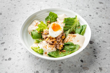 Fresh salad with romaine lettuce, creamy dressing, tuna, corn, soft-boiled egg, and crispy onions in a white bowl on a speckled countertop, offering a nutritious and delicious meal.