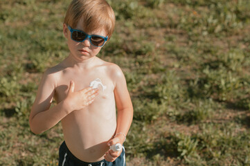 Little white-skinned boy in sunglasses applying sunscreen lotion on his body under bright sunlight. Concept of sun protection, summer, skin care, and child health outdoors.