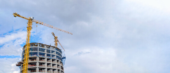 Construction cranes towering beside sleek skyscraper, working against cloudy urban skyline, representing contemporary architectural development and infrastructure growth © Newleks