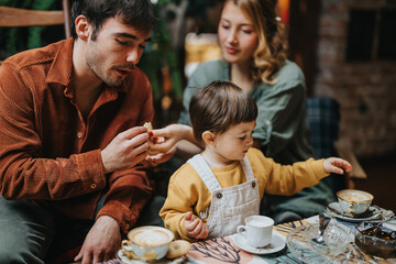 A family of parents and a child enjoying a warm tea party together indoors, engaging in an intimate...